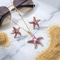 Parure étoile de mer or et émail violet lilas sur marbre blanc avec sable doré et lunettes de soleil